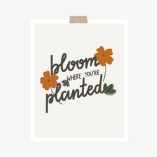 Load image into Gallery viewer, An art print featuring the popular quote, “Bloom where you’re planted,” written in calligraphy and decorated with orange flowers and green leaves hung with a piece of kraft tape on the top.
