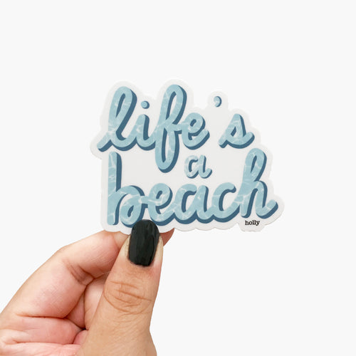 A funny pun sticker with blue text that reads, Life’s a beach, written in modern calligraphy with white water effects in the letters.