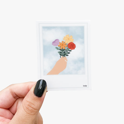 A sticker featuring a polaroid of a hand holding summer flowers and facing them up toward the sky filled with clouds.