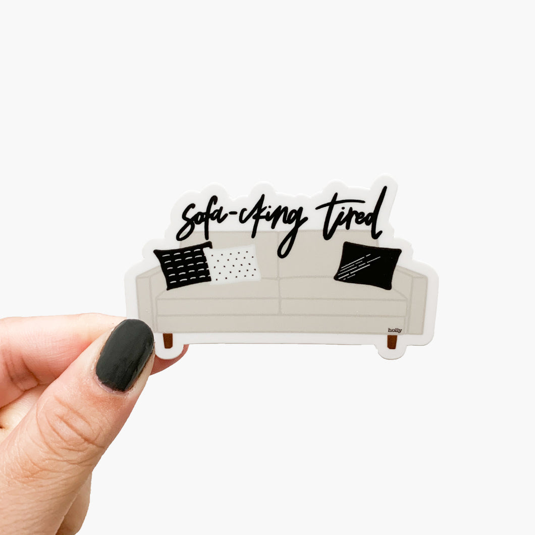 A funny sticker featuring a beige sofa with black and white decorative pillows and text that reads, Sofa-cking tired.