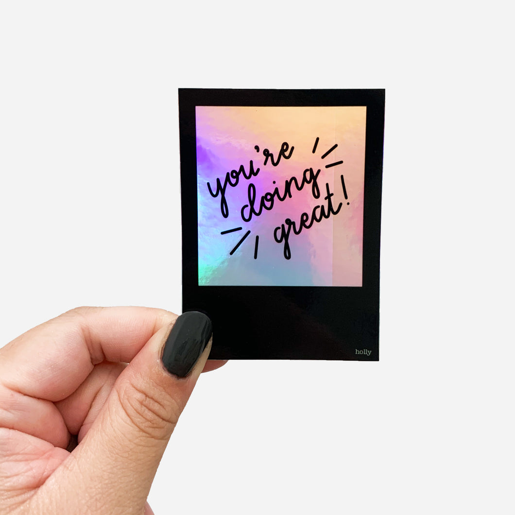 A sticker featuring a black polaroid with a holographic center and text that reads, You’re doing great!